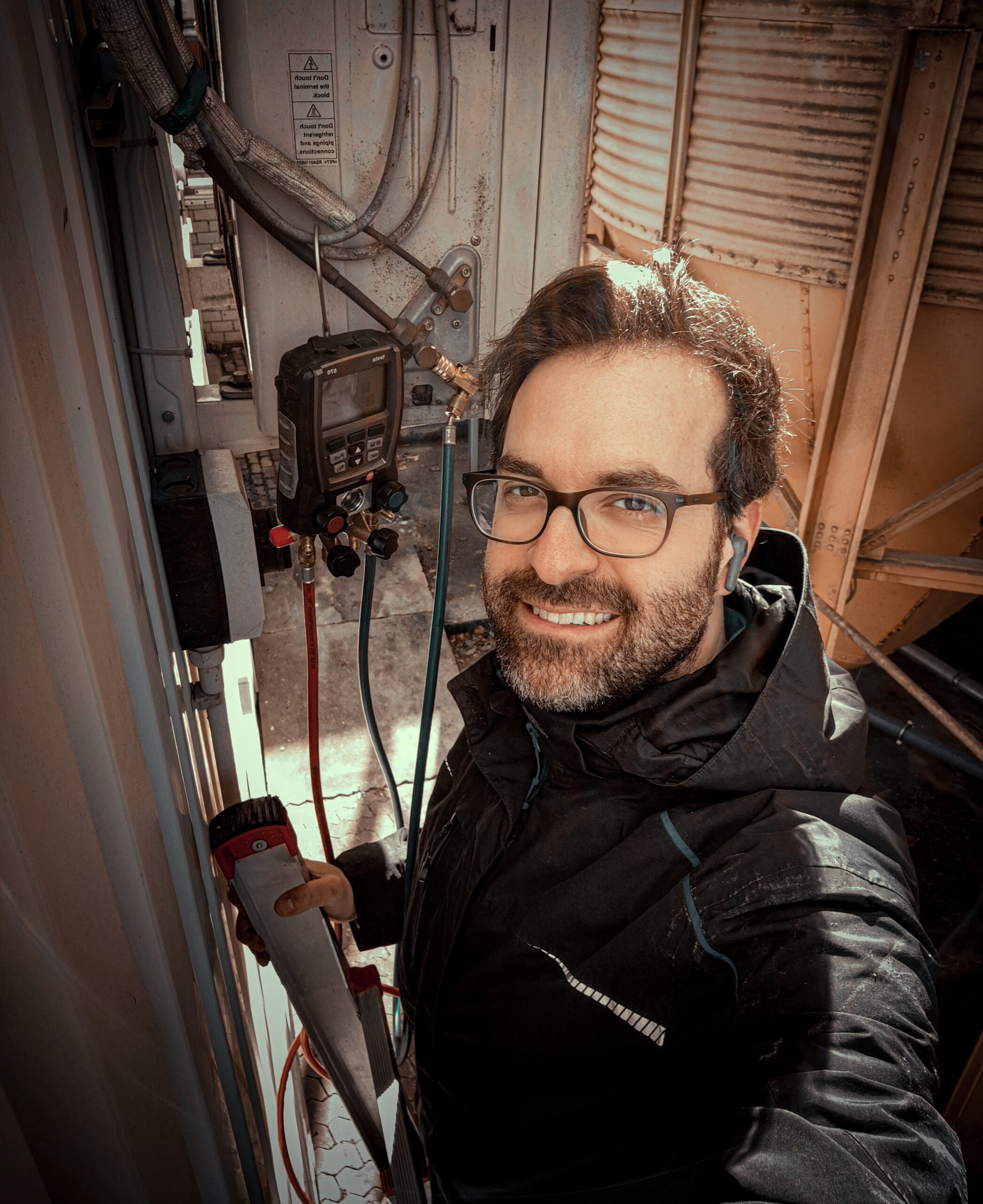 Ein Junger hübscher Mann mittleren Alters. Er steht auf einer Leiter. Grinsend sieht er in die Kamera und freut sich, dass heute so ein schöner Tag ist. IFRA Kältetechnik - Stefan Luber